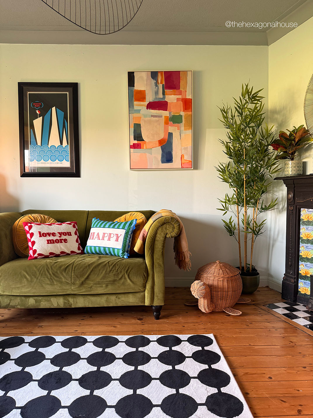 A styled living room corner featuring an olive green velvet sofa with colorful throw pillows, a black-and-white patterned rug, a tall potted bamboo plant, and abstract wall art on a light green wall.