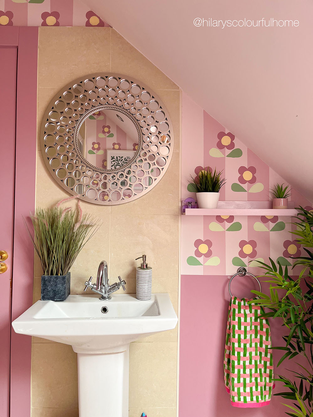 A styled pink bathroom corner featuring a white pedestal sink, a decorative circular mirror, potted plants, a pink shelf, and a green-and-white checkered towel.