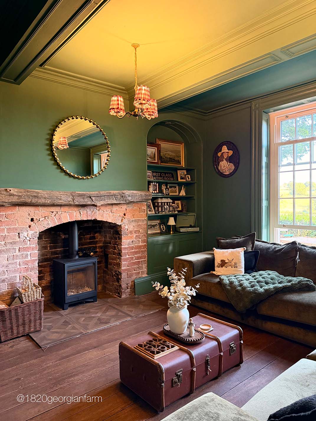 A cozy, rustic living room featuring a green wall, a brick fireplace with a wood-burning stove, a dark green sofa with a green quilt, a vintage brown trunk coffee table, and a warm yellow-gold ceiling.