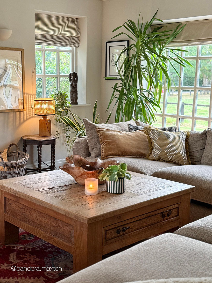 A rustic wooden coffee table in a cozy, sunlit living room with a beige sectional sofa, decorative pillows, a lit candle, and a small potted plant on top.