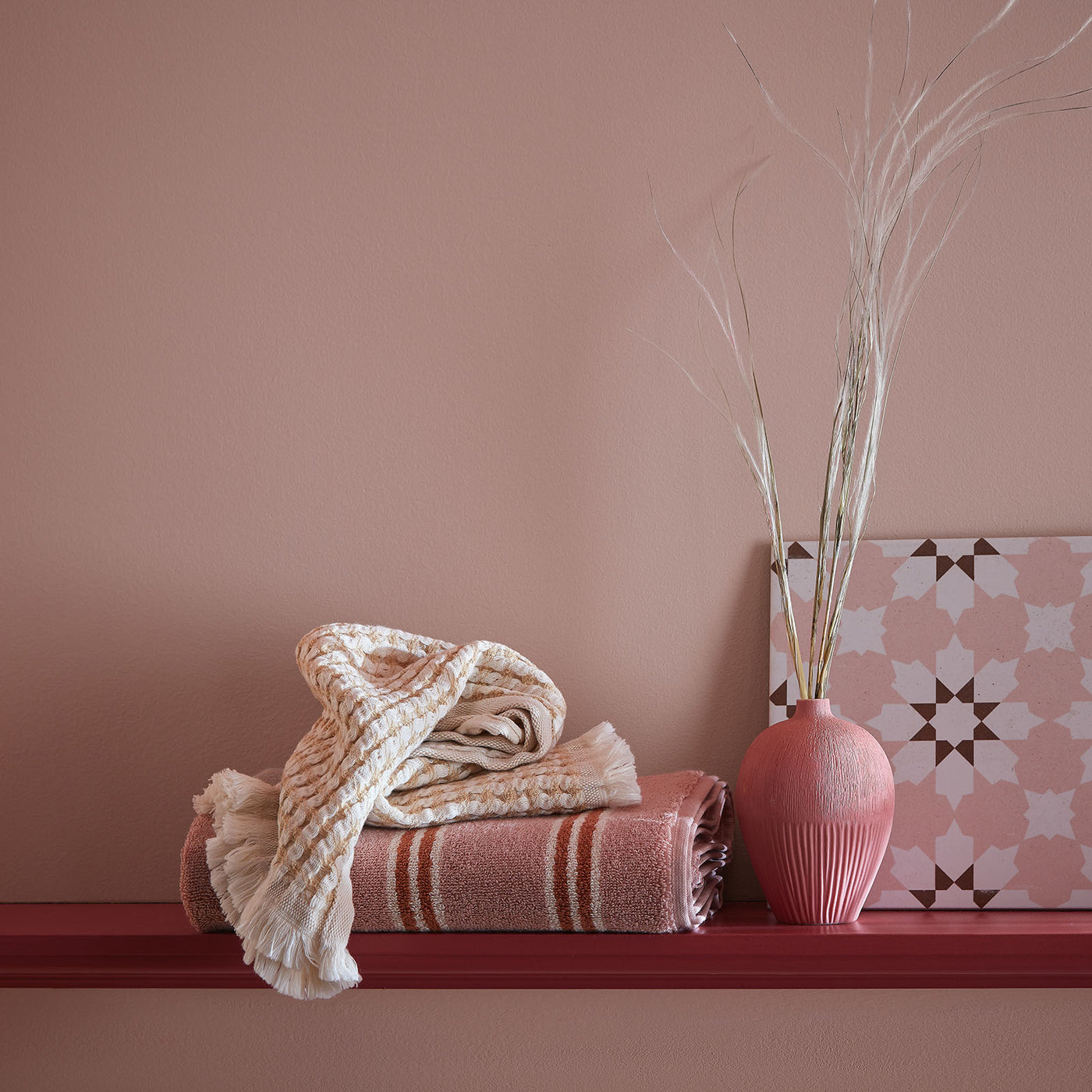 Stack of folded towels on a shelf with a pink vase and decorative item against a pink wall.