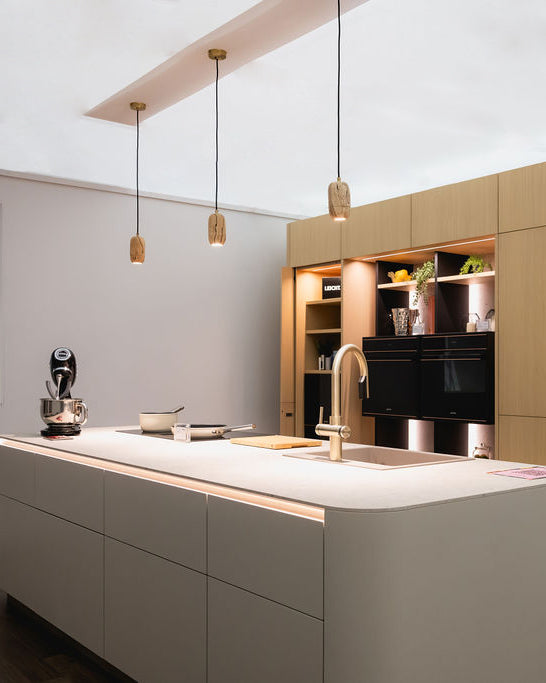 A modern kitchen interior with a large white island countertop, matte light gray lower cabinets, built-in black appliances, warm wood cabinetry, and three hanging pendant lights.