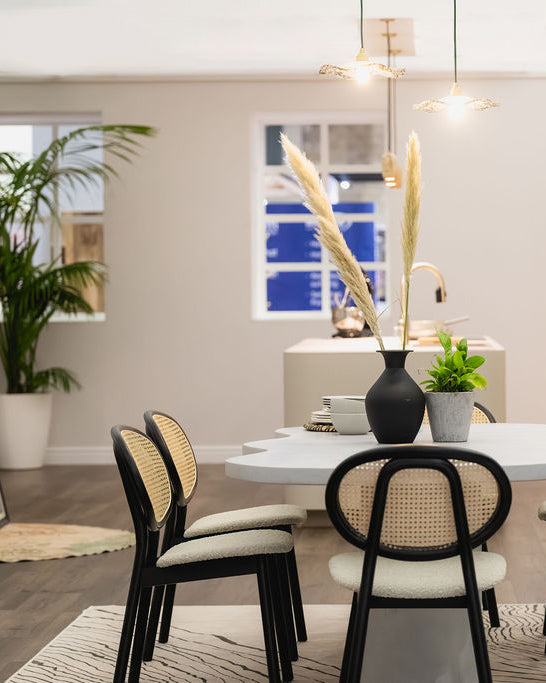 A modern dining area featuring a white oval table with black-framed chairs that have woven cane backs and cream upholstered seats. A tall black vase with pampas grass and a small green plant sit on the table, set against a neutral dining room background with a kitchen island and pendant lights.