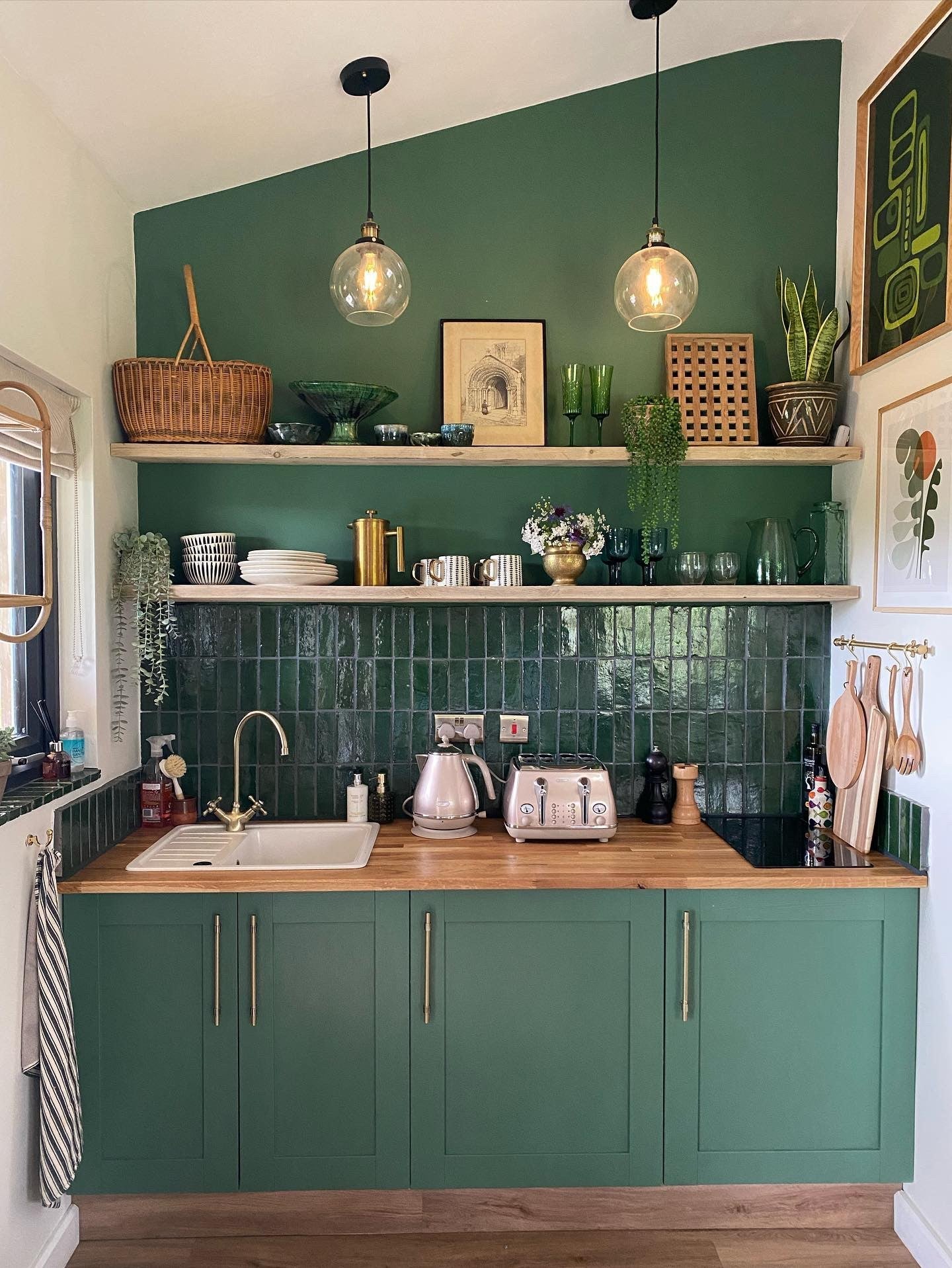 White and green kitchen nook with a sink and storage.|Split image of two different angles of a kitchen.|Open shelves with glasses, dishes and plants above a kitchen counter.|A kitchen with open plan shelving and a red oven with a round woven mat on the fl
