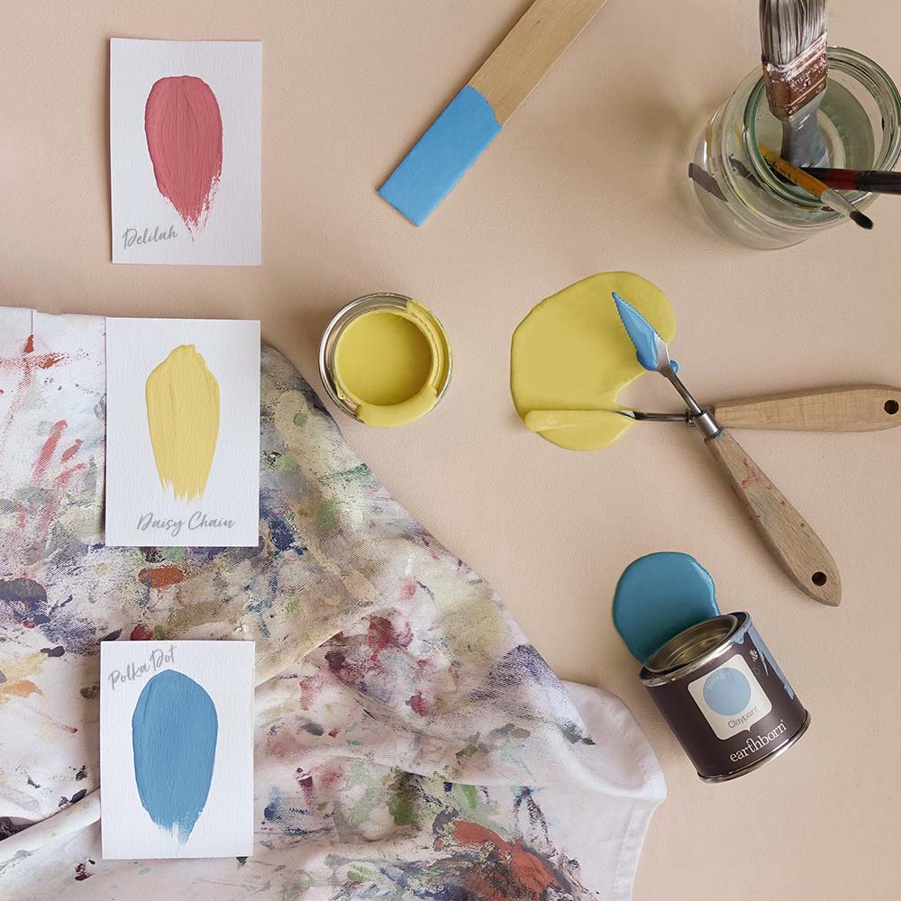 Lifestyle Arty family mood board.|Paper swatches on a well used apron with sample pots.|A bold striped background featuring a sample pot of paint.|A pink background showing Earthborn paint brushes.|A room decorated using Peach Baby on the walls.|Wall mura