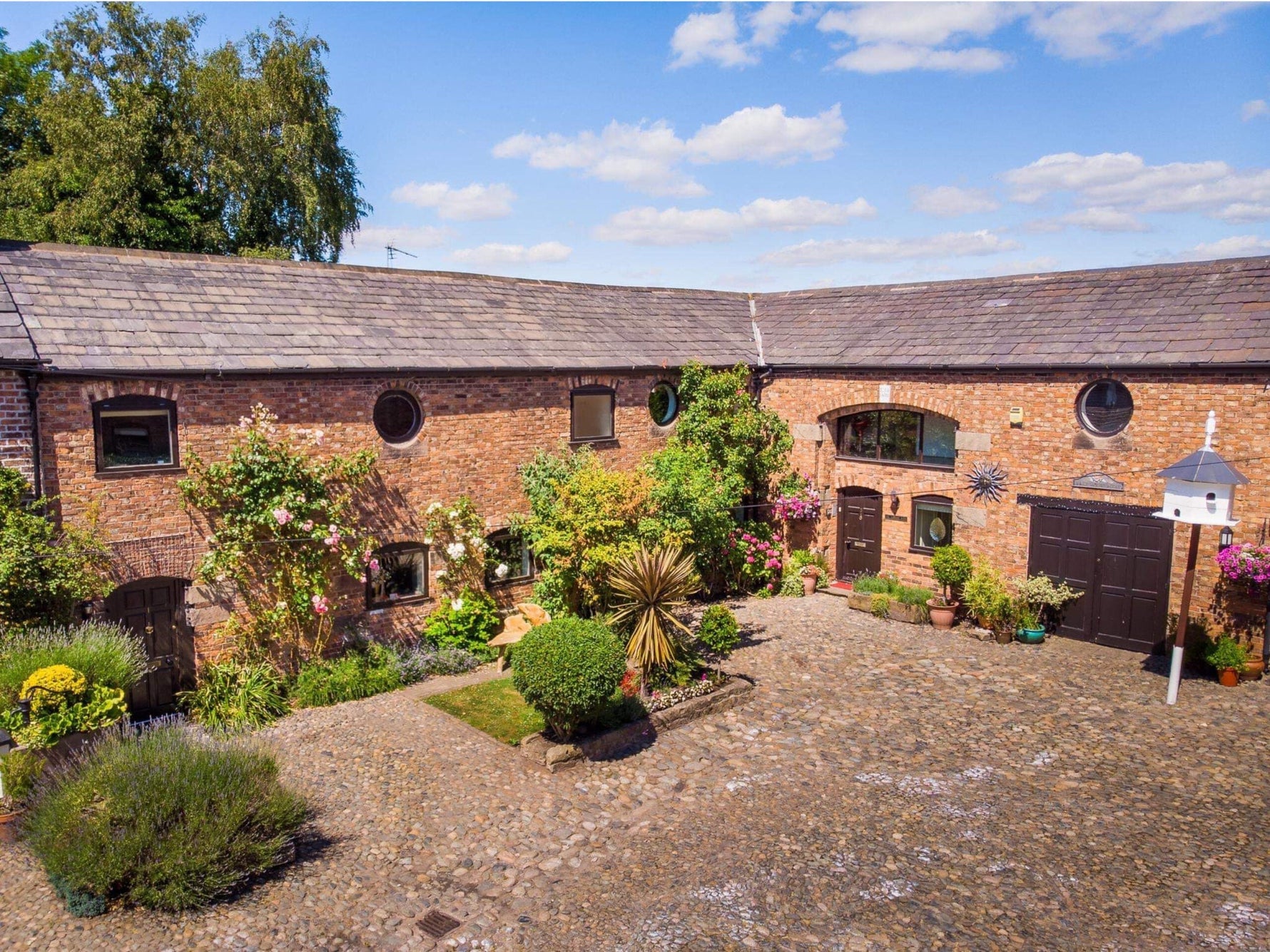 A beautiful barn conversion with a lovely spread of plants and flowers.|A raw brick fireplace with a tartan armchair and large window.|Bright orange SMEG fridge against a white wall with a small shelf above with colourful jugs.|Raw brick edging around the
