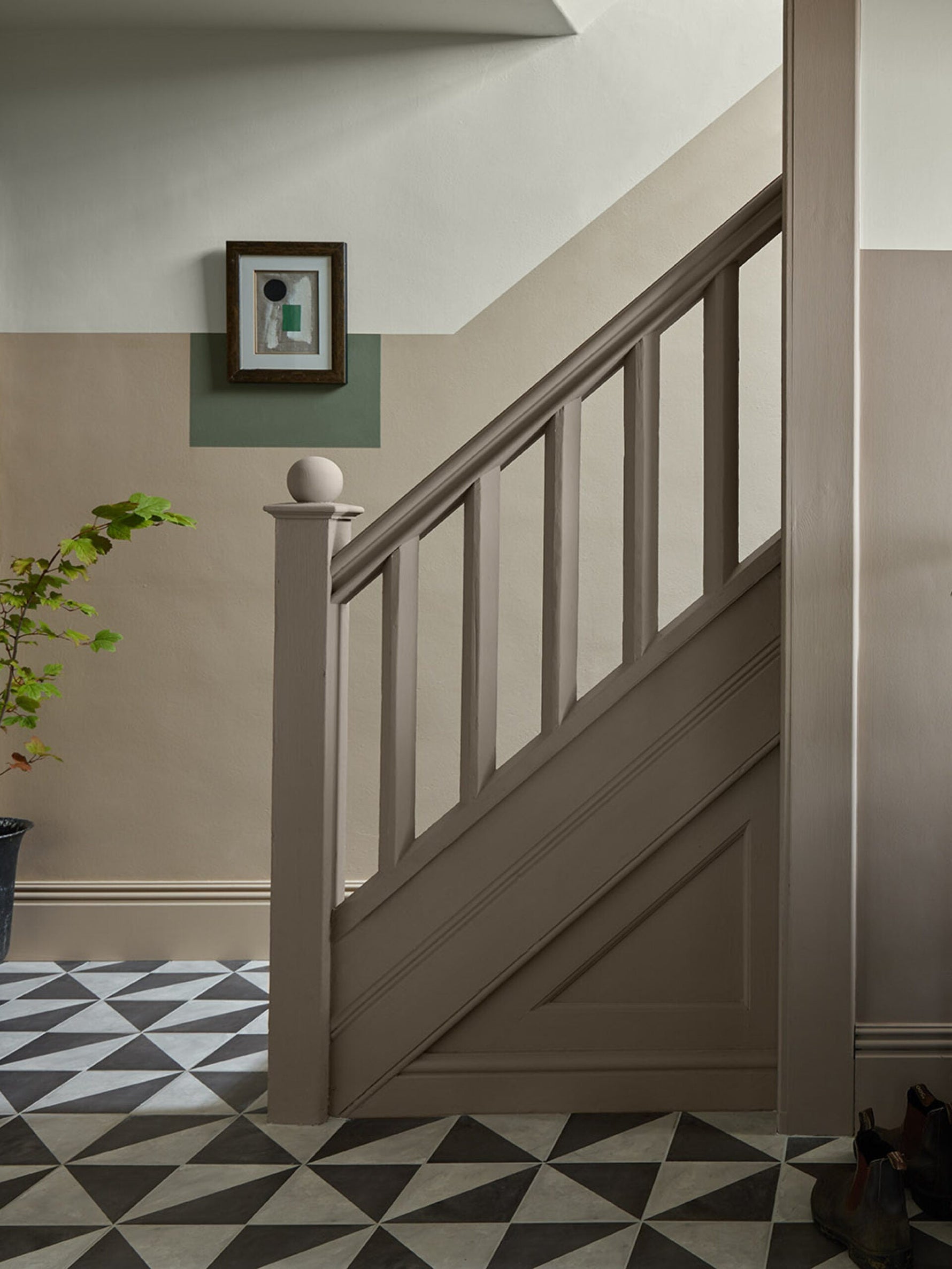 A patterned floor with a short banister leading upstairs. Colour blocking painted on the walls to add to the art hanging.|A striped wall with a table and a jug full of fresh yellow tulips.|A carefully crafted wooden stair case with a cute little black dog