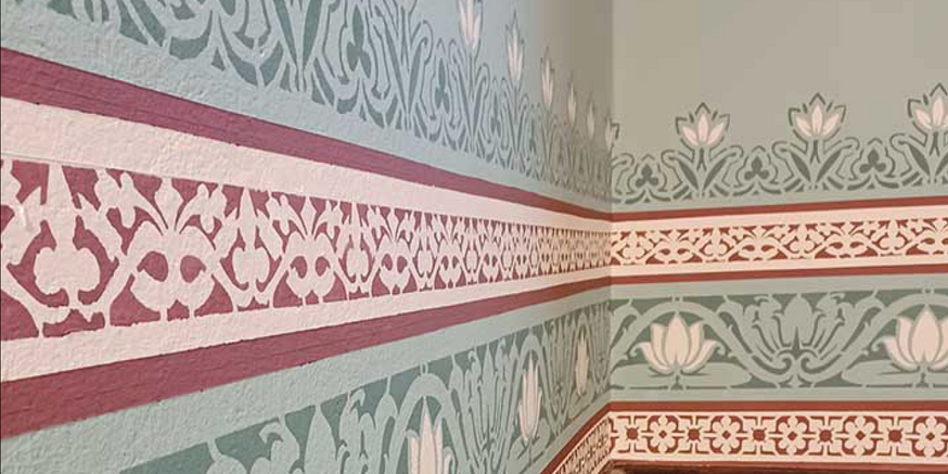 The corner of a wall with bold patterned edging and floral design.|Painted stamp print on a wall adding boarders to a plain wall behind a headboard.|A white wall with a patterned green trim around the boarders and door frame.|A wall with a checked boarder