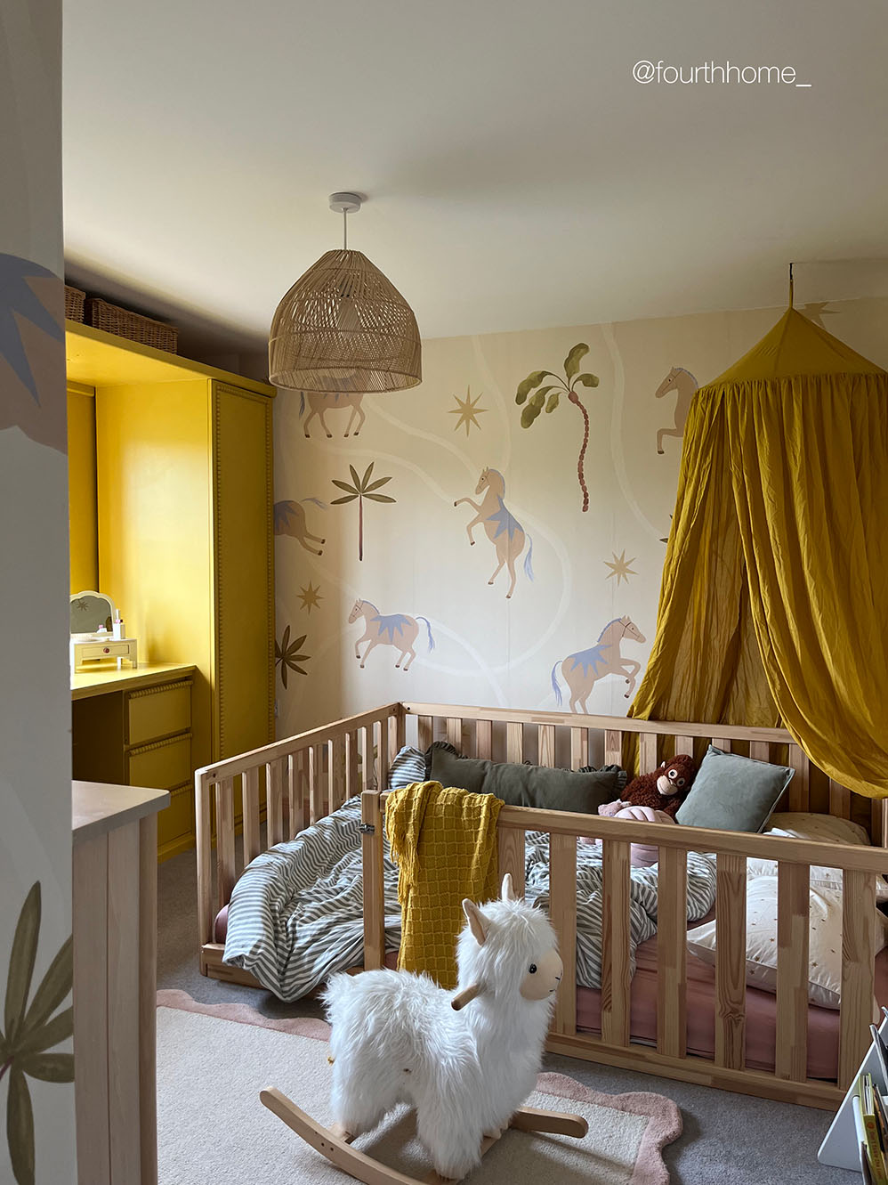 A styled children's bedroom featuring a light wood crib with bedding, a yellow canopy, a wicker pendant light, and a white llama toy in the foreground. The wall has a warm cream background with painted horses and palm trees.