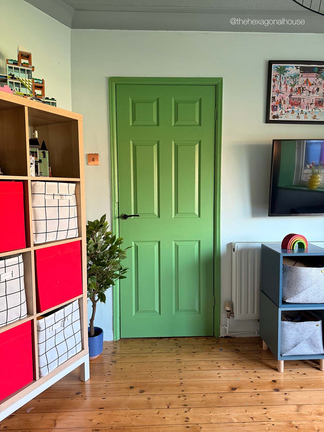 A bright green six-panel interior door set in a light green wall, shown in a styled room with a wooden floor, a shelving unit on the left, and a blue cabinet on the right.