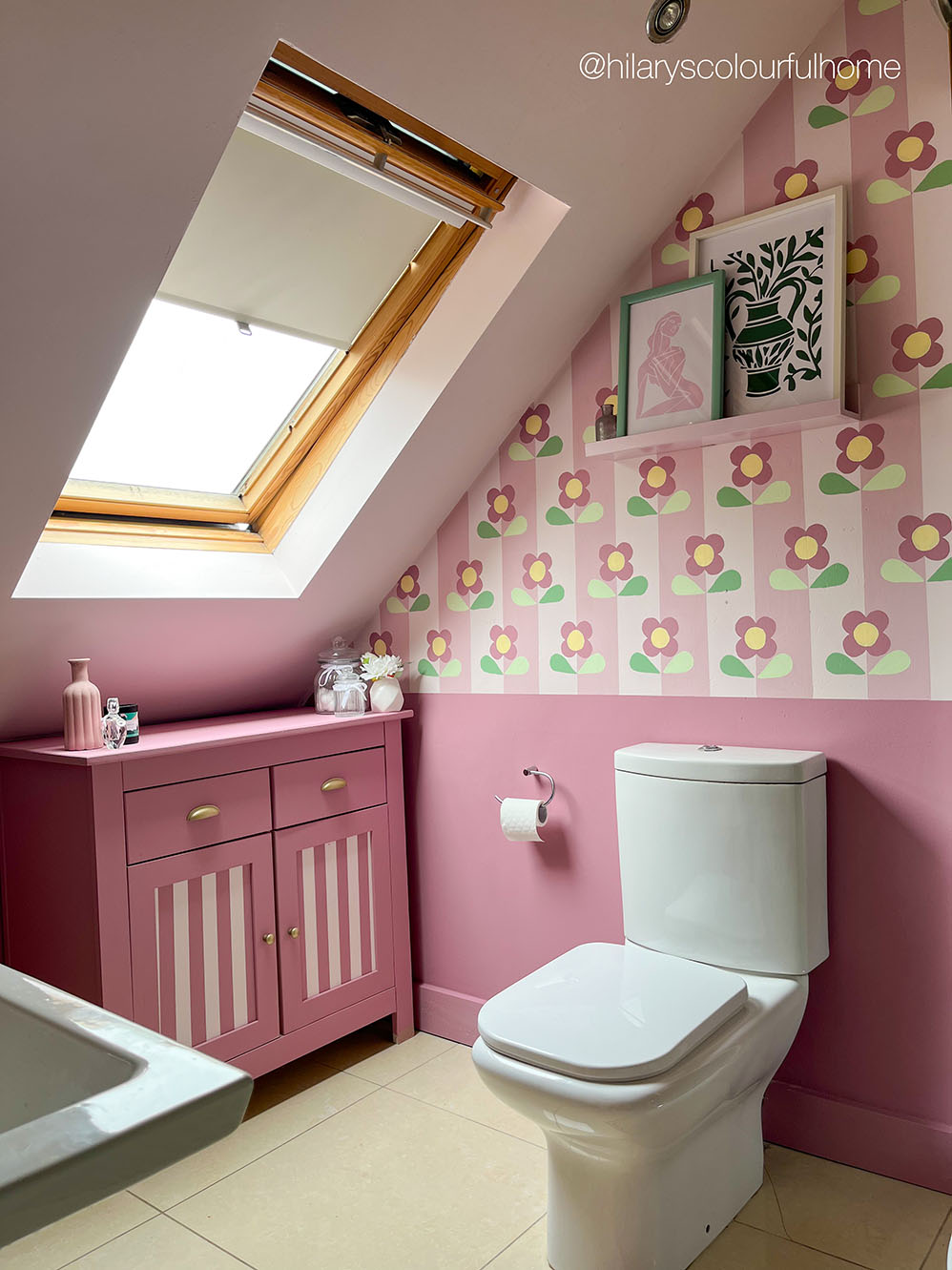 A styled pink bathroom with a floral painted wall, a pink vanity cabinet, a white toilet, and a skylight window.