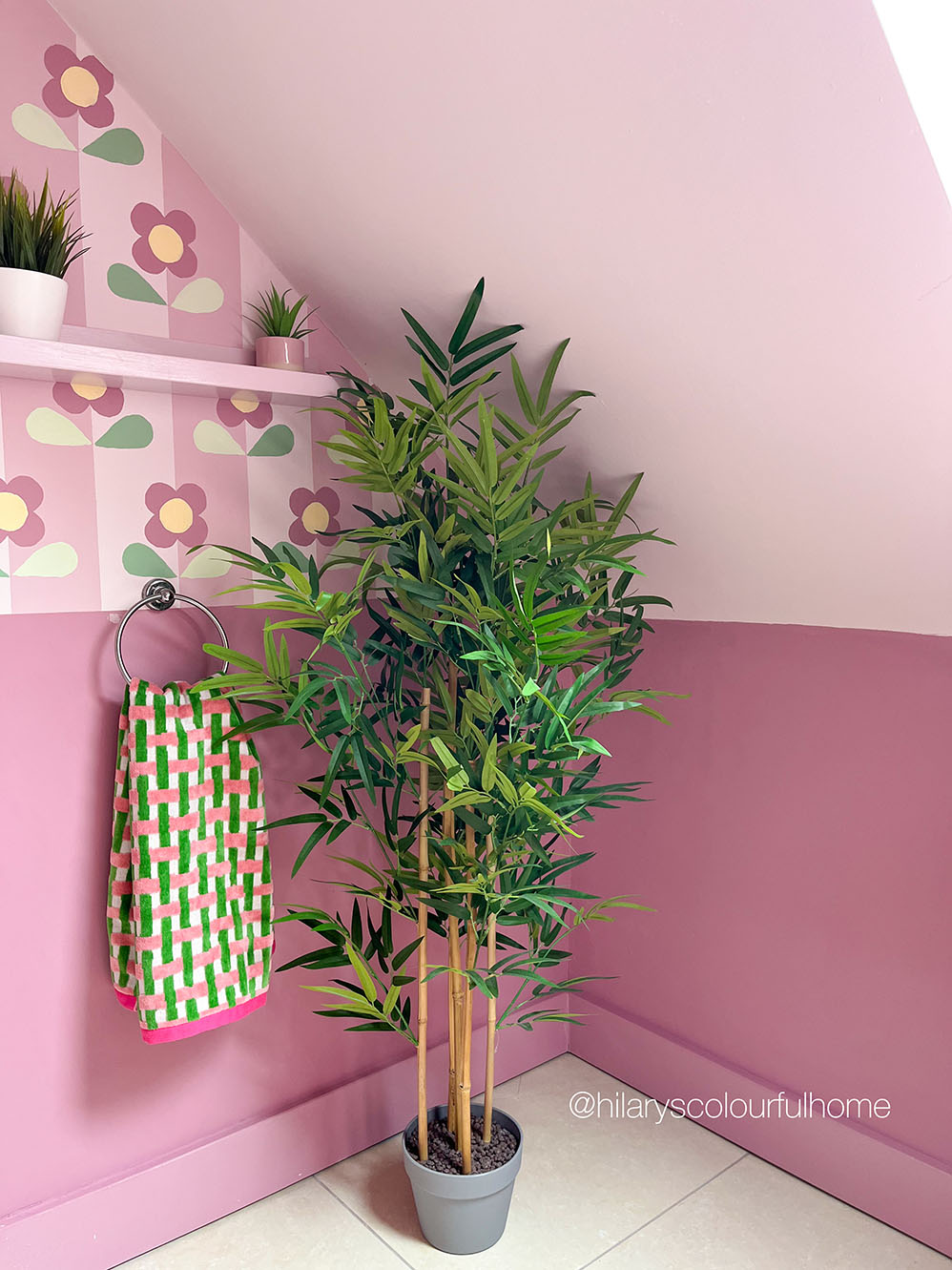 A tall artificial bamboo plant in a gray pot placed in the corner of a bathroom with pink walls, a shelf, and a hanging towel.