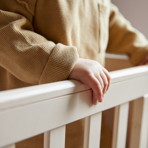A close-up of a child wearing a textured tan waffle-knit long-sleeve top, resting their hand on a white wooden crib