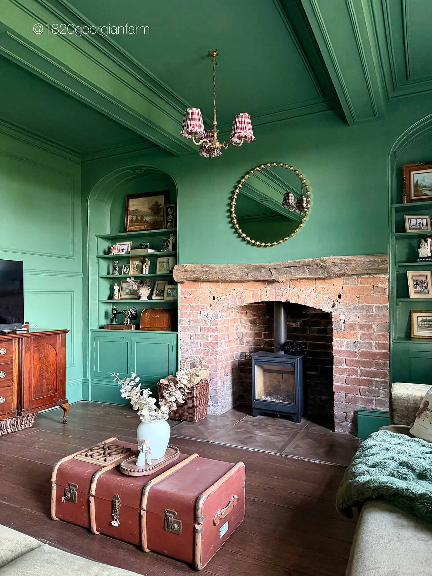 A styled living room with deep green walls and ceiling, featuring a brick fireplace with a wood-burning stove, a round gold-framed mirror, built-in shelves, and a red vintage suitcase used as a coffee table.