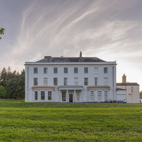 A large white classical-style manor house with a gray slate roof, situated on a green lawn under a cloudy sky with dramatic streaks of light.