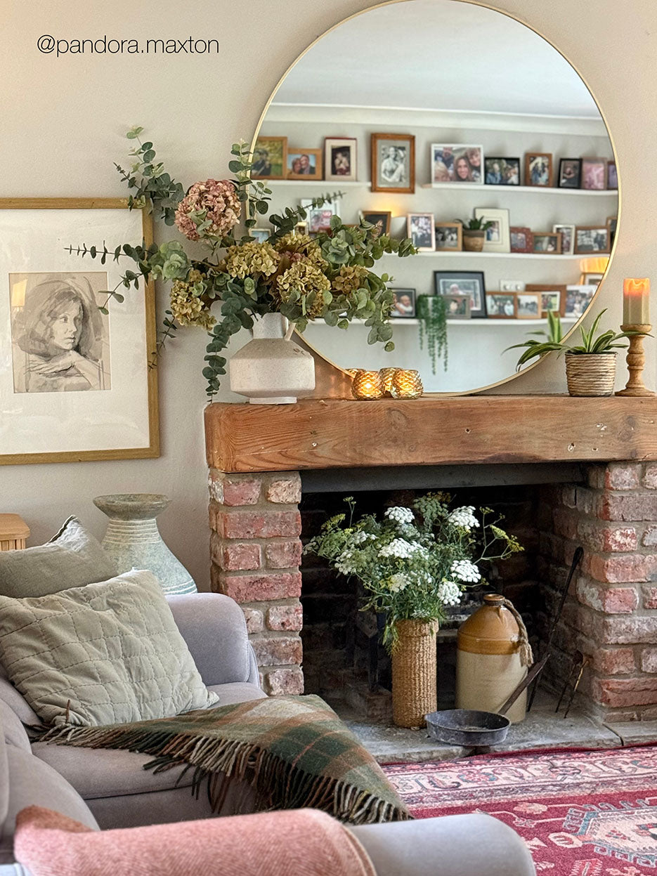 A cozy living room scene featuring a large round wall mirror with a gold frame above a rustic brick fireplace mantel decorated with dried flowers, greenery, and candles. A light gray sofa with a green plaid throw blanket is visible in the foreground, along with a patterned rug and a framed sketch on the wall.