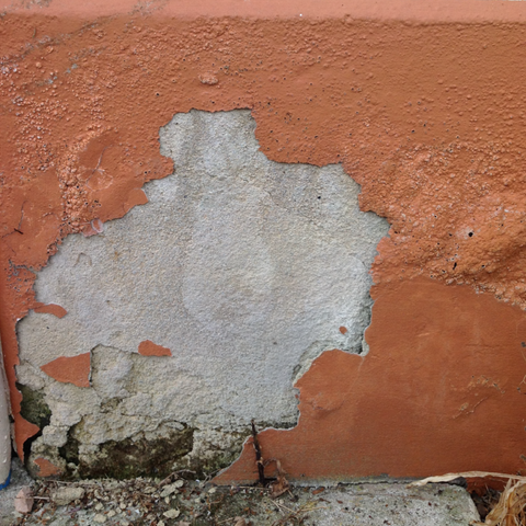 A close-up of a textured terracotta orange stucco wall with a large section of the paint peeled away to reveal a rough, light gray concrete or plaster surface underneath.