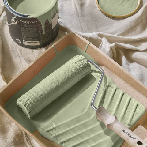 A paint roller covered in light sage green paint rests in a cardboard tray filled with matching paint, with a paint can and an open paint lid nearby on draped fabric.
