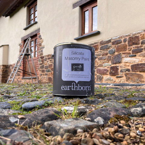 A can of earthborn Silicate Masonry Paint stands on a cobblestone ground in front of a stone and plaster exterior wall of a building.