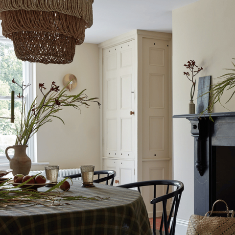 A styled dining room corner featuring a round table with a green and beige plaid tablecloth, black spindle-back chairs, a large woven wicker pendant light, and a tall cream cabinet in the background.