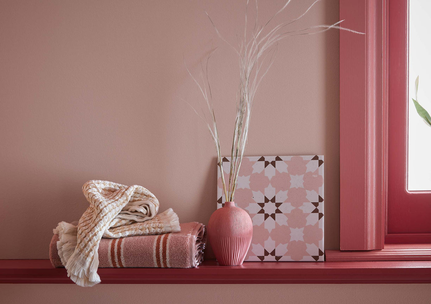 Decorative items on a windowsill including a pink vase, patterned box, and striped blanket against a pink wall.