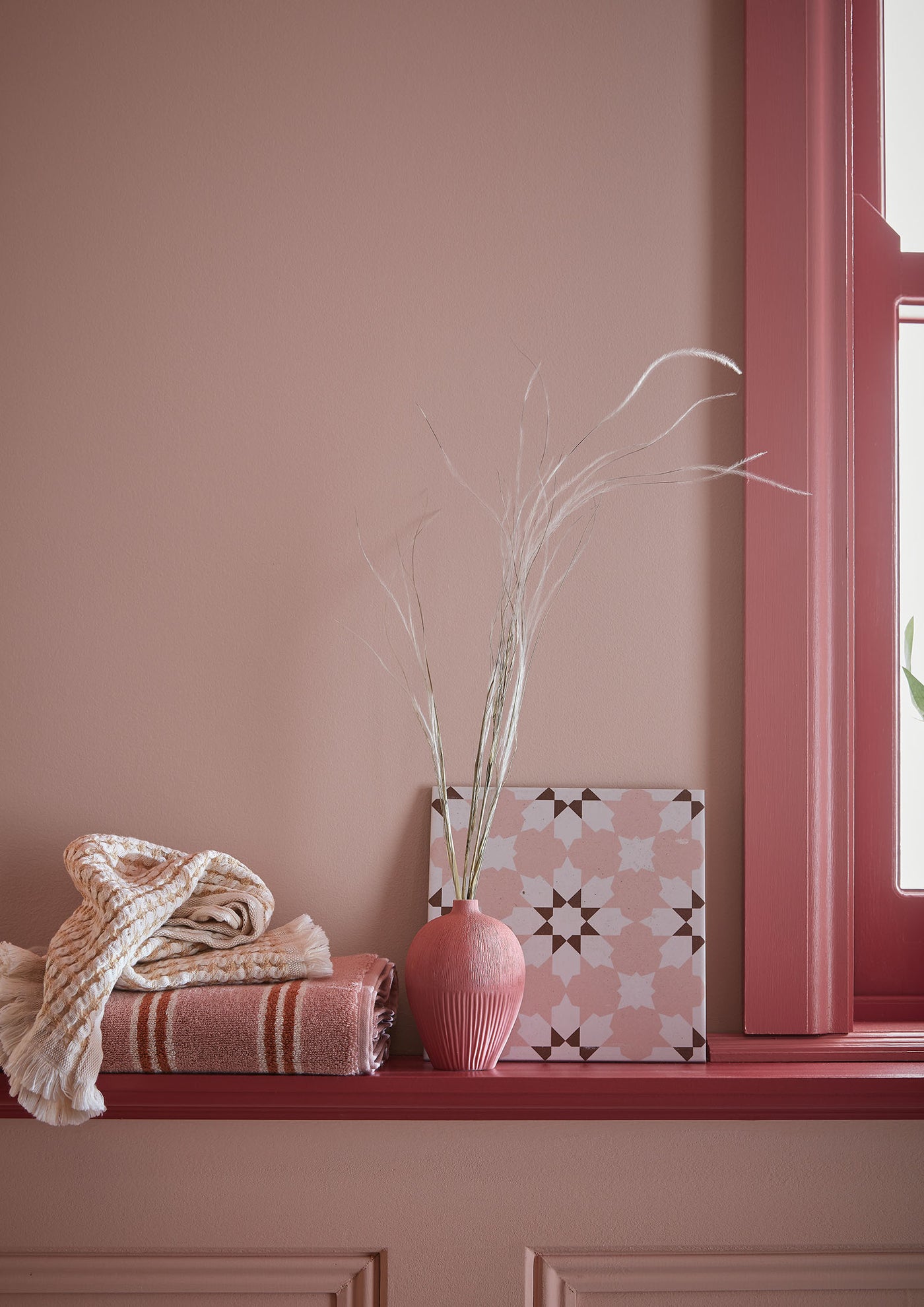 Pink window sill with a vase, blanket, and decorative box against a pink wall.