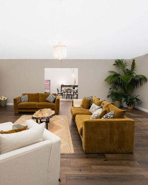 A modern living room featuring a prominent mustard yellow velvet sectional sofa with patterned throw pillows, a matching yellow loveseat in the background, a white armchair in the foreground, and a large potted palm plant.