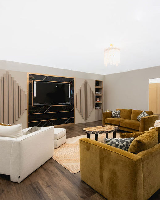A modern living room featuring a mustard yellow velvet sofa in the foreground, a white armchair on the left, and a matching mustard yellow sofa in the background. The room has dark wood floors, a black marble TV wall with gold trim, and decorative slatted wall panels.