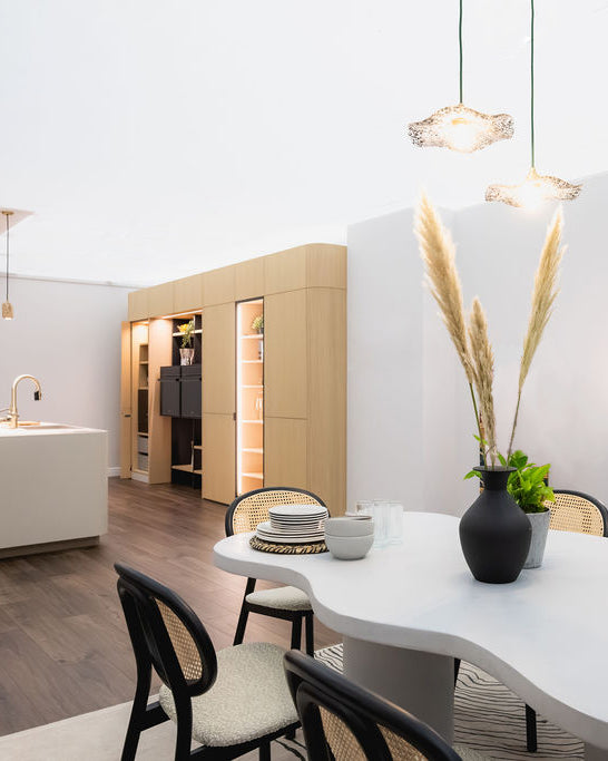 A modern dining area featuring a white organic-shaped table, woven-back chairs, a black ceramic vase with pampas grass, and a built-in light wood cabinet in the background.