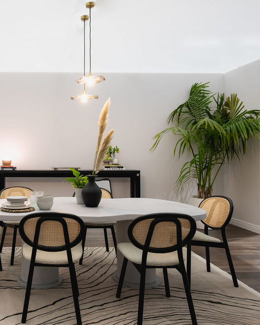 A modern dining room setup featuring a round white table with black-framed woven cane chairs, set on a patterned rug in a bright room with a potted palm and pendant lights.