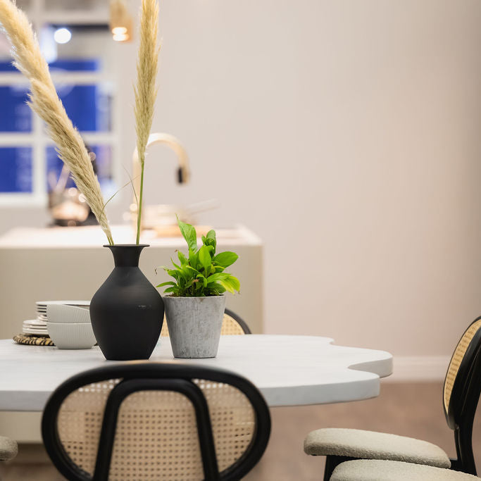 A styled dining table scene with a matte black vase holding tall beige pampas grass and a small green potted plant in a gray pot, set in a softly lit interior with chairs and a blurred kitchen background.