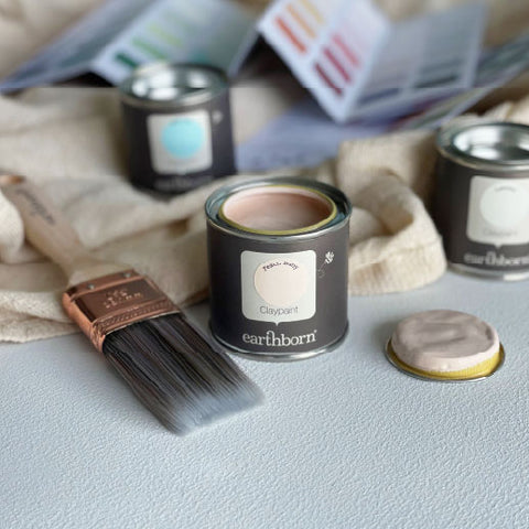 An open can of earthborn Claypoint clay paint sits on a light gray surface, with a paintbrush, a small lid, and other paint cans and color swatch cards blurred in the background.