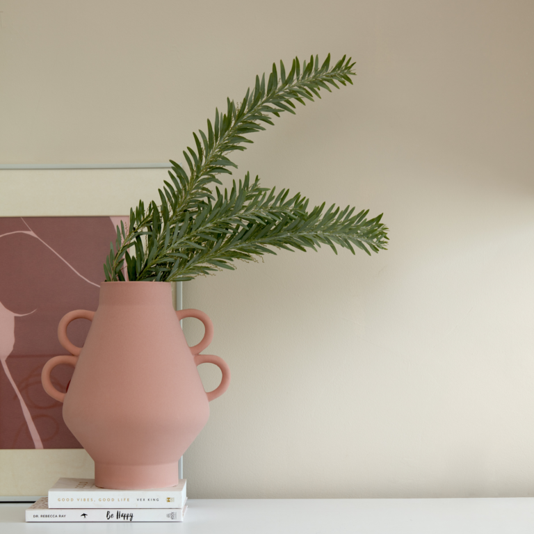 A pink vase balancing on two books with green leaves sticking out of it with an abstract frames painting in the background.